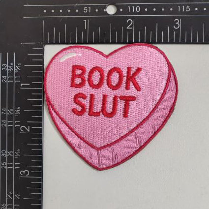 Heart-shaped patch with 'BOOK SLUT' text on a white background with ruler for scale.